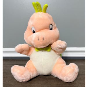 May include: A plush toy dragon in a seated position. The dragon is primarily peach-colored with a white belly and a green bow tie. It has large, round eyes and green, horn-like features on its head. The toy is soft and cuddly.