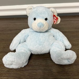 May include: A light blue plush teddy bear with a white muzzle and inner ears. The bear has black eyes and a red heart-shaped tag attached to its ear. The bear is sitting on a wooden surface.