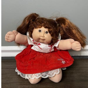 May include: A vintage doll with brown hair styled in pigtails. The doll has a round face, large eyes, and is wearing a red velvet dress with white lace trim. The doll's arms and legs are soft and plush.