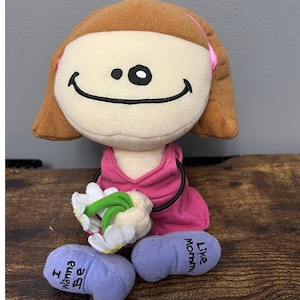 Puede incluir: Una niña de peluche con cabello castaño, vestida con un vestido rosa y zapatos azules. Lleva un ramo de flores blancas con tallos verdes. El texto en sus zapatos dice "I Wanna Be Like Mommy".