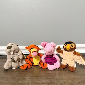 Könnte beinhalten: Ein Set aus vier Plüschtieren, bestehend aus einem grauen Biber, einem orange-schwarz gestreiften Tiger, einem rosa Ferkel in einem dunkelviolett gestreiften Hemd und einer braun-cremefarbenen Eule mit gelben Augen und Schnabel, in einer Reihe.