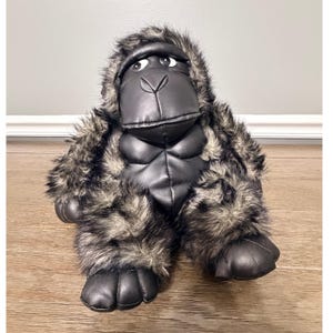 Puede incluir: Un peluche de gorila con cara, pecho y manos de piel sintética negra lisa, contrastado con pelo sintético largo y desgreñado de color gris y negro que cubre los brazos, las piernas y la cabeza. El gorila tiene ojos grandes y está sentado.