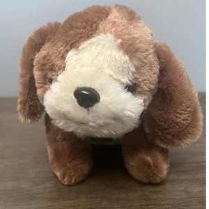 May include: A plush toy dog with brown and white fur. The dog has floppy ears, a black nose, and a friendly expression. The toy is standing on a wooden surface.