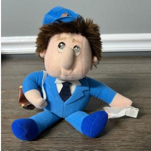 May include: A blue plush toy of a mail carrier wearing a blue uniform, a blue hat, and a brown bag. The toy has brown hair and a large nose.