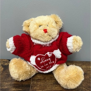 May include: A plush teddy bear with tan fur and a red sweater. The sweater has white trim and a heart-shaped patch with the phrase "Love is in the air". The bear has a red nose and is sitting on a wooden surface.