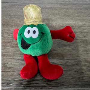 Puede incluir: Un personaje de peluche con cuerpo verde, brazos y piernas rojos y sombrero dorado. El personaje tiene una cara sonriente con ojos blancos y boca negra. El juguete está diseñado para estar de pie.