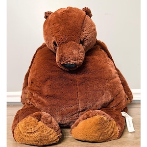 May include: A large, plush brown teddy bear with a soft, furry texture. The bear has a dark brown nose and lighter brown paws. It is sitting upright, with its arms and legs extended. A white tag is attached to the bear's leg.