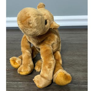 May include: A tan plush camel stuffed animal with soft fur, a rounded snout, small ears, and four legs with darker tan accents on the feet. The toy features a smooth, velvety texture and a seated posture with a visible hump on its back.
