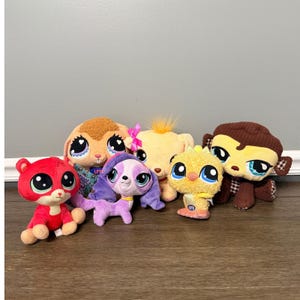 以下が含まれることがあります： 赤、茶色、紫、黄色など、さまざまな色のぬいぐるみ動物のおもちゃのコレクション。おもちゃは大きく表現力豊かな目を持ち、木製の表面に配置されています。おもちゃは、さまざまな動物に似るように設計されています。