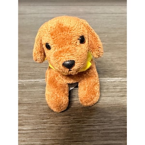 May include: A plush toy dog with brown fur, black eyes, and a black nose. The dog has a yellow collar and is sitting on a wooden surface. The toy is a small, cuddly representation of a dog.