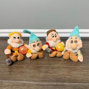 May include: Four plush dwarf dolls with white beards and hats. The first wears a yellow hat and orange outfit with an axe. The second wears a light blue hat labeled BASHFUL. The third wears a brown hat and red shirt. The fourth wears a light blue hat labeled SLEEPY.