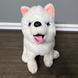 May include: A plush white dog toy with blue eyes, a black nose, and a pink tongue. The dog has pink inner ears and is sitting upright. The toy is made of soft, fluffy material.