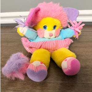 May include: A vintage plush toy with a yellow face, pink fur, and blue eyes. It has pink and purple ears, blue arms, and a pink and white striped skirt. The toy's feet are yellow with pink and white striped bottoms.