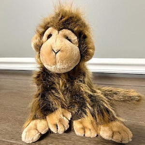 May include: A plush monkey toy featuring long, shaggy brown and tan fur. The stuffed animal has a light tan face with a soft, rounded muzzle, dark brown eyes, and small ears. It is depicted in a seated position with visible front paws and a tail.