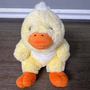 Puede incluir: Un pato de peluche amarillo con patas y pico naranja. El pato tiene un mechón de pelo blanco en la cabeza.