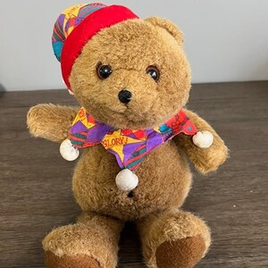 May include: A brown teddy bear wearing a red and white hat with a multi-colored scarf that says "Glory!"