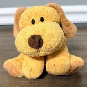 Puede incluir: Un perro de peluche con cuerpo marrón claro, orejas marrones oscuras y nariz marrón. El perro tiene ojos negros y está sentado sobre una superficie de madera. El juguete es suave y tierno.
