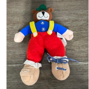 May include: A plush bear doll wearing a blue shirt, red pants, and tan shoes. The bear has a brown head with a green hat and yellow suspenders. The doll has white lace trim on the shoes.