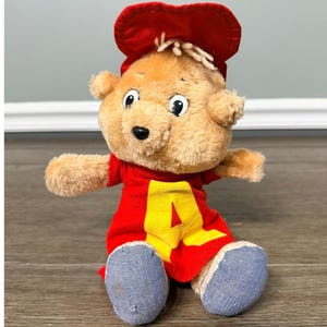 Puede incluir: Un peluche de un personaje de oso de dibujos animados. El oso tiene pelaje marrón claro, ojos negros y una nariz negra. Lleva una camisa roja con una "A" amarilla en la parte delantera, un sombrero rojo y pantalones vaqueros azules. El juguete está sentado.