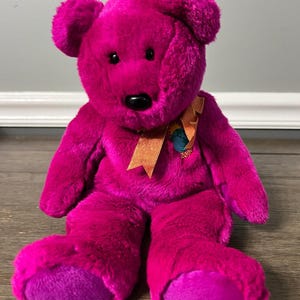 May include: A large, plush pink teddy bear with a purple nose and orange ribbon. The bear is sitting on a wooden floor and has a tag on its foot that says "2003".