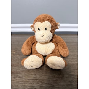 以下が含まれることがあります： 茶色の毛皮とクリーム色の顔、胸、足を持つぬいぐるみのおもちゃの猿。ぬいぐるみには黒いボタンの目と小さな耳が付いています。猿は木製の表面に座っています。