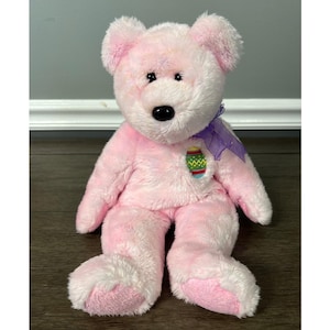May include: Pink and white plush Easter bear with a purple ribbon and an embroidered Easter egg on its chest.