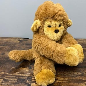 May include: A plush brown gorilla toy with a light brown face, ears, and feet. The toy has a soft, furry texture and is sitting on a wooden surface. The gorilla has a small, dark brown nose and black eyes.