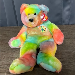 Könnte beinhalten: Ein bunter Batik-Plüschbär mit einem Peace-Zeichen auf der Brust. Der Bär sitzt auf einer braunen Oberfläche. Der Bär hat ein rotes Etikett mit einem weißen Stern.
