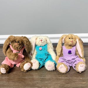 May include: Three plush rabbit toys sitting in a row. The left rabbit is brown with pink overalls, the middle rabbit is cream with teal overalls and a pink rose, and the right rabbit is tan with lavender overalls and a purple rose.