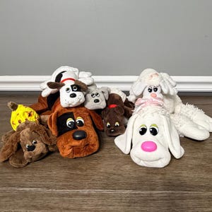 May include: A collection of plush puppy dog toys in various colors and sizes. The toys include brown, white, and yellow puppies with different facial expressions. Some have floppy ears and pink noses. The toys are arranged on a wooden surface.
