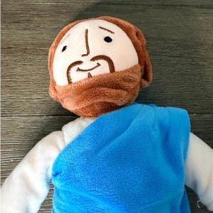 May include: A plush doll depicting a religious figure, featuring a light skin tone, brown hair and beard, and a blue robe. The doll has embroidered facial features and is made of soft, textured fabric. The background is a wooden surface.