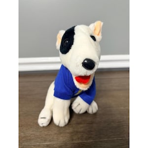 May include: A plush white and black Bull Terrier dog wearing a blue shirt with a yellow lightning bolt design. The dog has a red mouth and black eyes. The dog is sitting on a wooden surface.