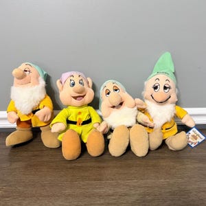Op de afbeelding: Vier pluche poppen die personages uit Sneeuwwitje en de zeven dwergen voorstellen. De poppen hebben zachte stoffen lichamen, gele tunieken, bruine schoenen en puntmutsen in lichtgroen of roze. Eén muts heeft de geborduurde naam Bashful.
