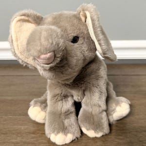 Könnte beinhalten: Ein kuscheliges, graues Elefanten-Spielzeug mit großen Ohren, einem Rüssel und kleinen schwarzen Augen. Der Elefant sitzt auf einer Holzoberfläche. Das Spielzeug hat eine weiche, pelzige Textur und eine hellrosa Nase.
