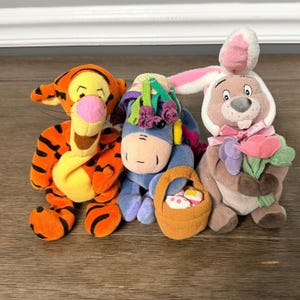 Könnte beinhalten: Drei Stofftiere: Tigger, Eeyore und ein Hase. Tigger ist orange mit schwarzen Streifen. Eeyore ist blau mit Strohhut und Korb. Der Hase ist braun und weiß und hält Blumen. Die Spielzeuge befinden sich auf einer Holzoberfläche.
