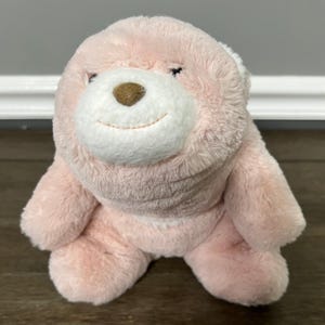 Op de afbeelding: Een roze teddybeer met een witte snuit en een bruine neus. De beer heeft een lachende uitdrukking en zit rechtop. De vacht is zacht en pluizig, waardoor hij er knuffelig uitziet.