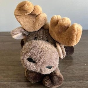 May include: A plush moose toy featuring a dark brown fuzzy body, a lighter tan snout, and large, soft, tan-colored antlers. The stuffed animal has small black eyes and a black nose, with a textured, soft fabric finish throughout its entire form.