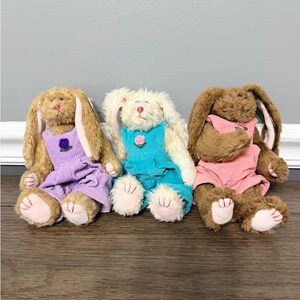 May include: Three plush stuffed rabbits with long ears and pink paw pads. The left rabbit is tan wearing lavender corduroy overalls with a purple flower. The center rabbit is white wearing teal corduroy overalls with a pink flower. The right rabbit is brown wearing pink corduroy overalls with a small flower.
