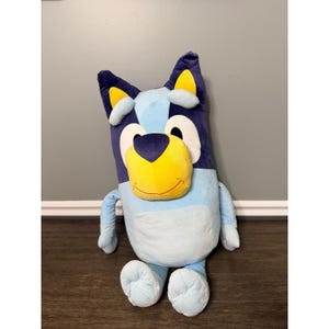 May include: A plush toy character featuring a light blue body with dark blue patches on the ears and head. The character has a yellow muzzle, a dark blue nose, and large white eyes. It has yellow inner ears and a light blue belly patch.