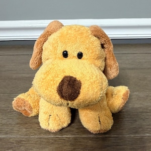 May include: A plush dog toy with a light brown body, darker brown ears, and a brown nose. The dog has black button eyes and is sitting on a wooden surface. The toy is soft and cuddly, perfect for children.
