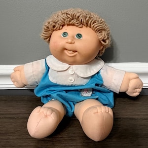 May include: A soft-bodied doll with light skin, green eyes, and curly tan yarn hair. The doll wears a white collared shirt with two buttons under a bright blue jumper. The doll has a rounded face, a small nose, and a single visible tooth in its mouth.