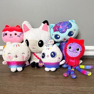 May include: A collection of six plush toys including a pink cat, a white llama, a blue mermaid cat with scales, two white cupcake-themed cats with colourful bases, and a purple cat wearing a red hoodie with long, multi-coloured striped tentacles.