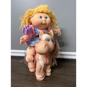 May include: A vintage Cabbage Patch Kids doll riding a tan and white horse toy. The doll has blonde yarn hair, blue eyes, and is wearing a floral dress. The horse has a pink bridle.