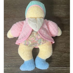 May include: A soft doll with a yellow body, wearing a pink jacket, a paisley patterned dress, and blue boots. The doll has a light blue bonnet and a white heart-shaped bib. The doll is made of a soft, plush material.