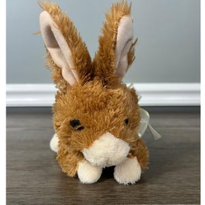 Puede incluir: Un juguete de peluche de conejo marrón con orejas largas y erguidas que presentan un forro interior beige claro. El animal de peluche tiene un pelaje marrón texturizado, hocico y patas de color crema, ojos de plástico oscuro y un lazo crema.