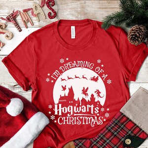 May include: Red t-shirt with a white graphic design that says "I'm dreaming of a Hogwarts Christmas". The graphic features a white silhouette of Hogwarts castle, a flying reindeer, and a flying sleigh.