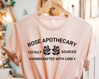 May include: A light pink t-shirt with the text "ROSE APOTHECARY" above two rose illustrations and the words "LOCALLY SOURCED HANDCRAFTED WITH CARE". The shirt is hanging on a wooden hanger.