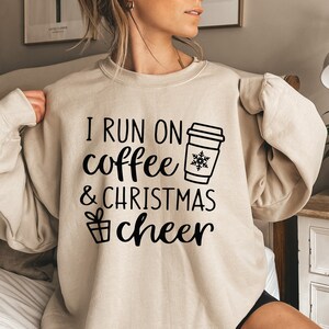 May include: A beige sweatshirt with the text "I run on coffee & christmas cheer" in black. A black and white illustration of a coffee cup with a snowflake is next to the text.