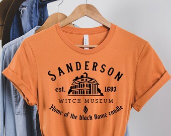 May include: Orange t-shirt with a black graphic design that says "Sanderson est. 1693 Witch Museum Home of the black flame candle". The graphic also includes a black silhouette of a house.