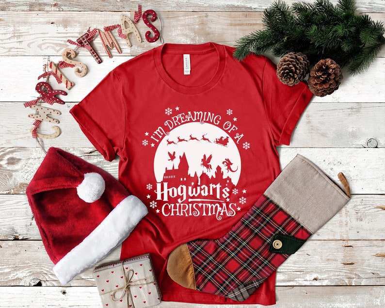 May include: Red t-shirt with a white graphic design that says "I'm dreaming of a Hogwarts Christmas". The graphic features a white silhouette of Hogwarts castle, a flying reindeer, and a flying sleigh.